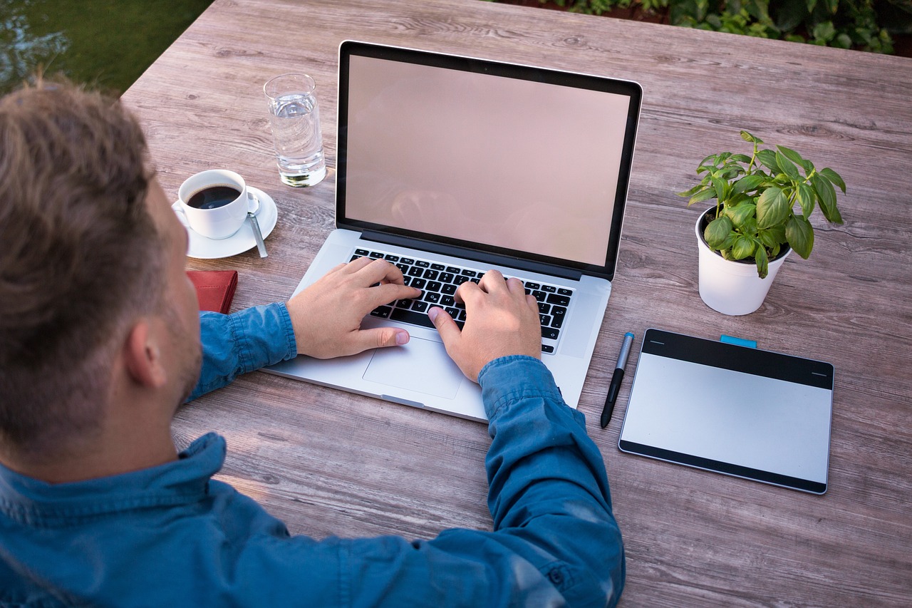 a man is working on his laptop, coffee, water, pen, diary and a plant on table
