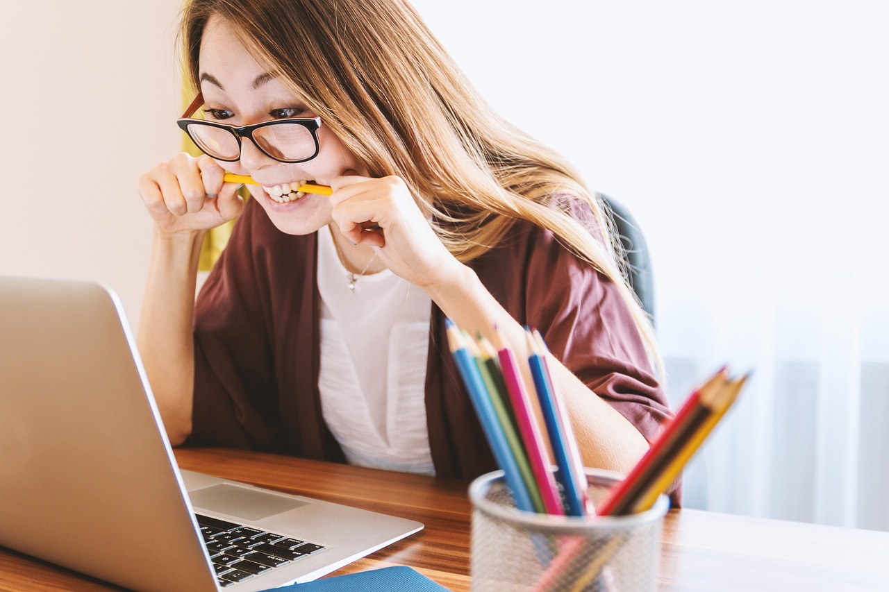 girl  is sitting on the desk, looking at the laptop and chewing pen